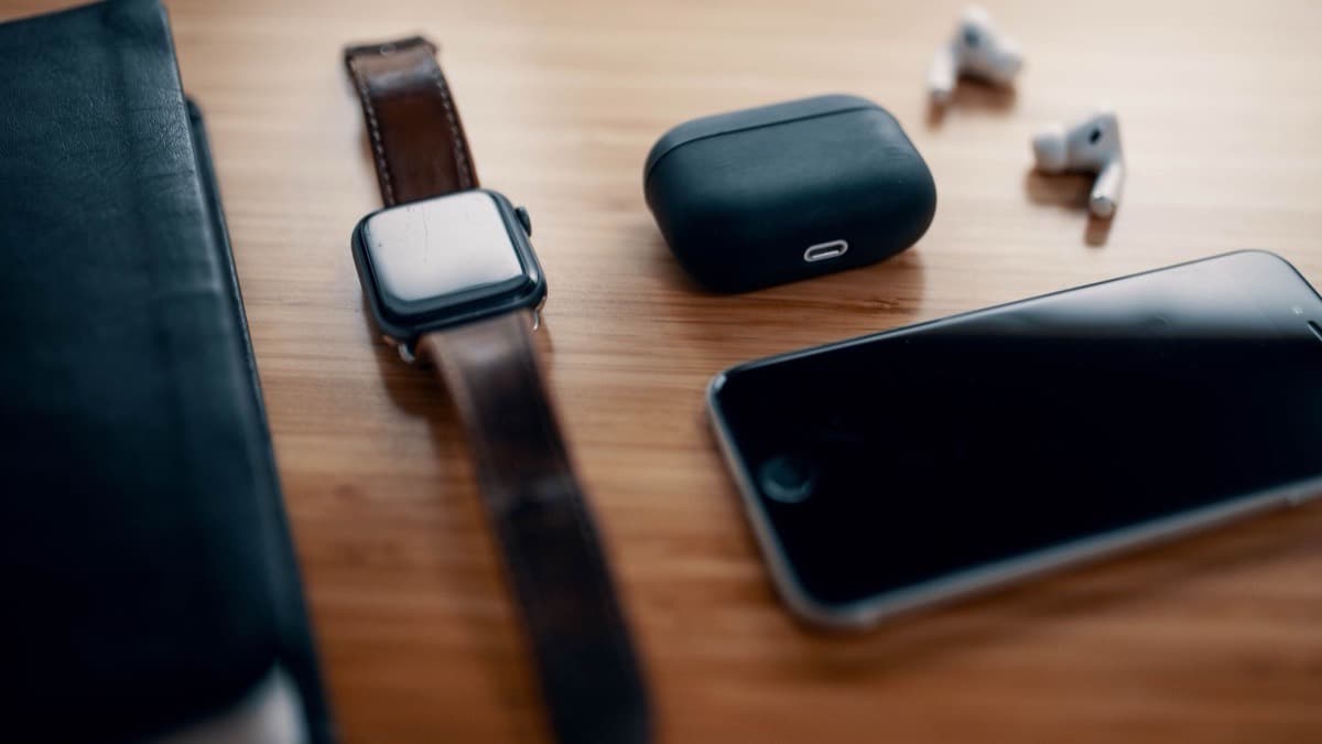 A used Apple Watch with a scratched screen and a worn leather strap on a wooden table, beside earbuds and an iPhone.