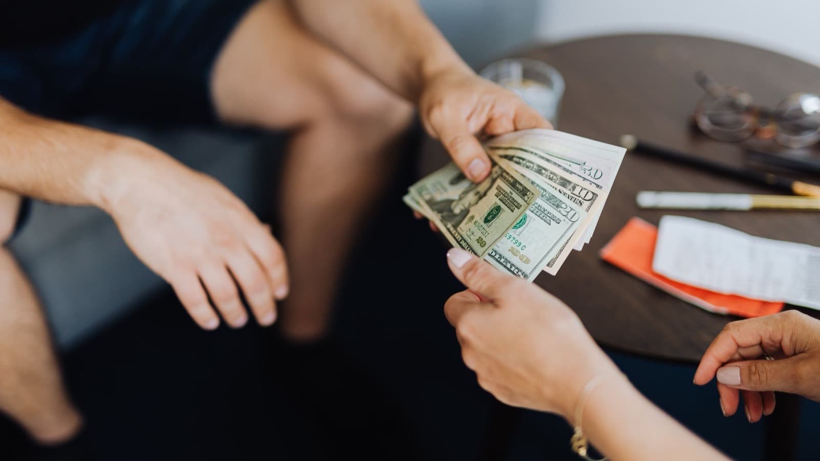 One person handing a fan of ten, twenty and fifty dollar bills into another person’s open hand across a small table.