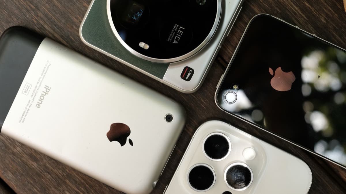 The original 2007 iPhone with its aluminium back, lying on dark wood beside two modern smartphones.