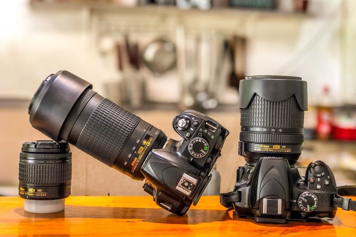Two Nikon DSLR camera bodies standing on a wooden kitchen table beside two spare zoom lenses.