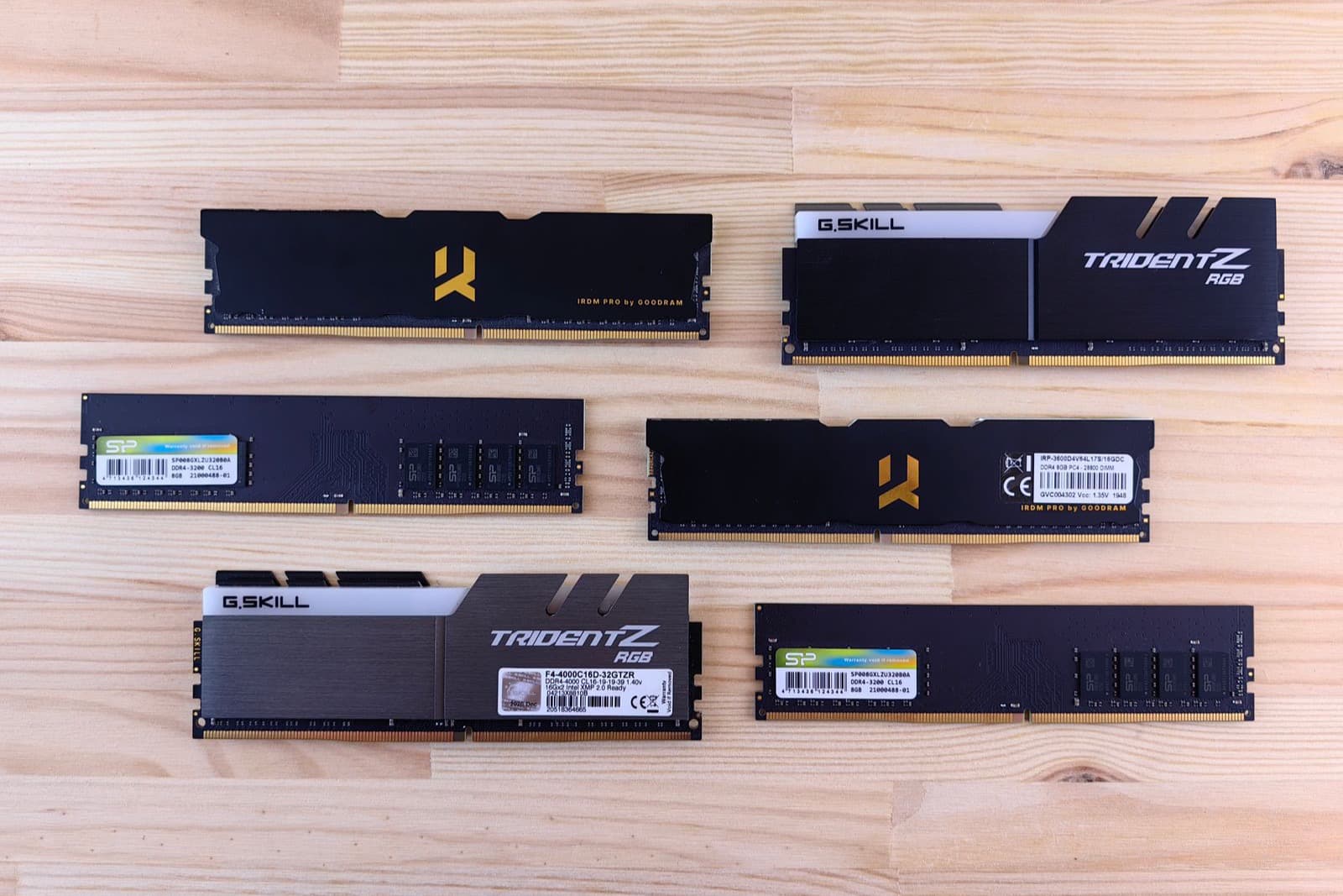 Six sticks of DDR4 desktop memory arranged in two columns on a pale pine table. Two have tall black and white heatsinks, two have low black heatsinks, and two are bare circuit boards with their specification labels facing up.