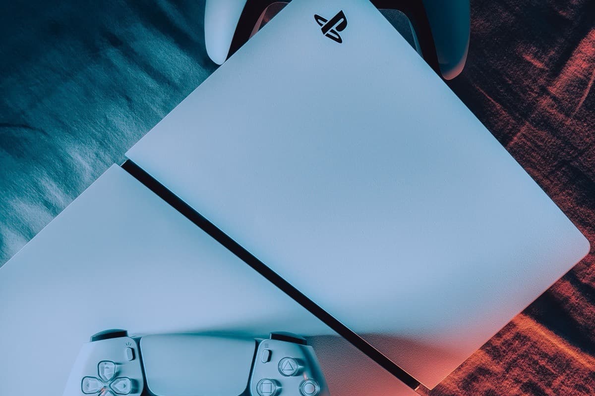A white PlayStation 5 console lying flat with two matching controllers, lit in teal and orange.
