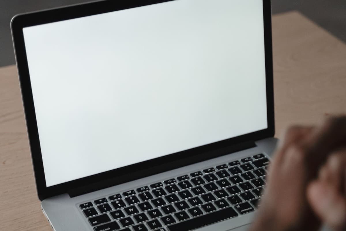 An open MacBook Pro with a blank screen on a pale wooden table, a person’s hands resting near the keyboard.