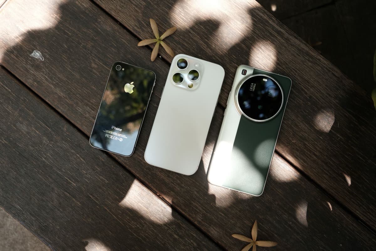 An older iPhone, a recent iPhone Pro and an Android phone laid in a row on a weathered wooden table in dappled sunlight.
