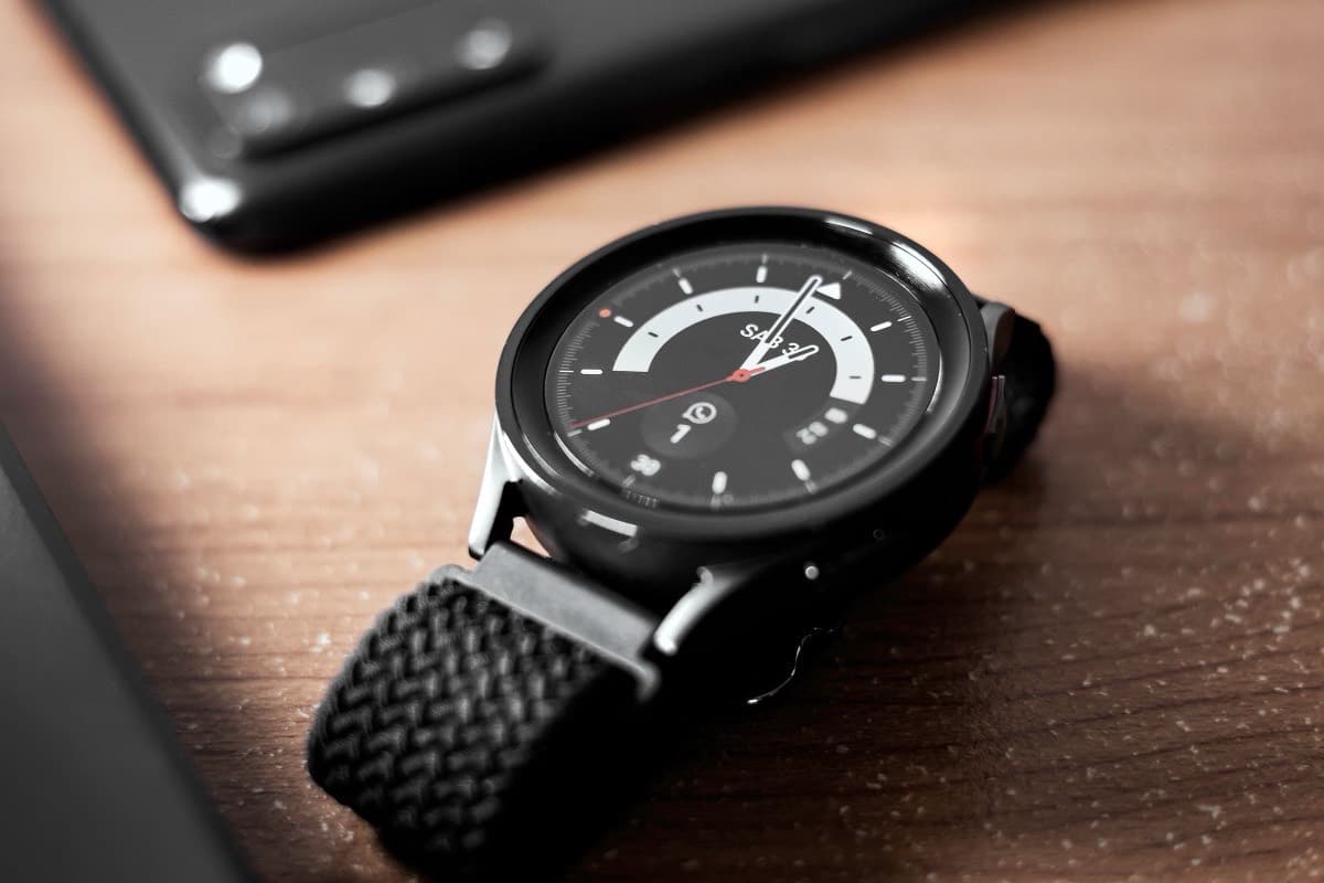 A round Samsung Galaxy Watch with a braided black strap lying on a wooden table beside a black Android phone.